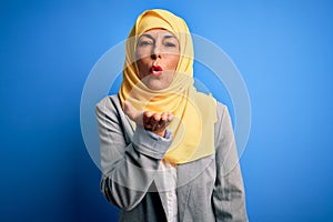 Middle age brunette business woman wearing muslim traditional hijab over blue background looking at the camera blowing a kiss with