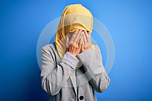 Middle age brunette business woman wearing muslim traditional hijab over blue background with sad expression covering face with