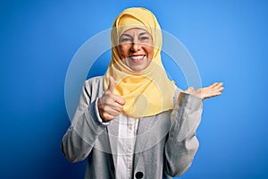 Middle age brunette business woman wearing muslim traditional hijab over blue background Showing palm hand and doing ok gesture