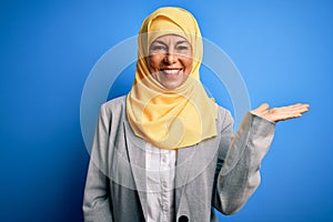 Middle age brunette business woman wearing muslim traditional hijab over blue background smiling cheerful presenting and pointing