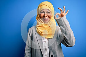 Middle age brunette business woman wearing muslim traditional hijab over blue background smiling positive doing ok sign with hand