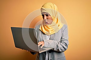 Middle age brunette business woman wearing traditional muslim hijab using laptop scared in shock with a surprise face, afraid and