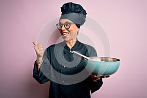 Middle age brunette chef woman wearing cooker uniform and hat using whisk and bowl pointing and showing with thumb up to the side