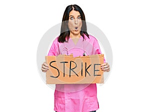 Middle age brunette doctor woman holding strike banner cardboard scared and amazed with open mouth for surprise, disbelief face