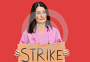 Middle age brunette doctor woman holding strike banner cardboard thinking attitude and sober expression looking self confident