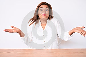 Middle age brunette hispanic business woman wearing casual white shirt sitting on the table clueless and confused expression with