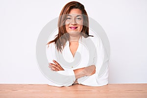 Middle age brunette hispanic business woman wearing casual white shirt sitting on the table happy face smiling with crossed arms