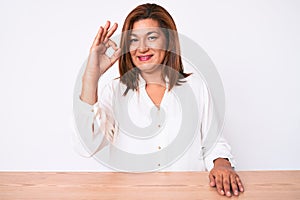 Middle age brunette hispanic business woman wearing casual white shirt sitting on the table smiling positive doing ok sign with