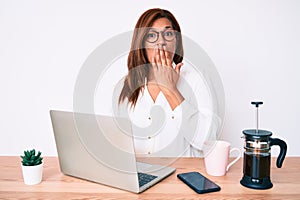 Middle age brunette hispanic business woman working at the office covering mouth with hand, shocked and afraid for mistake