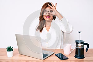 Middle age brunette hispanic business woman working at the office smiling happy doing ok sign with hand on eye looking through