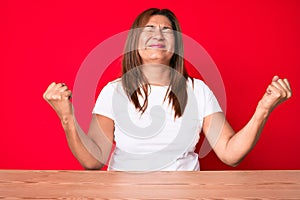 Middle age brunette hispanic woman wearing casual style sitting on the table very happy and excited doing winner gesture with arms