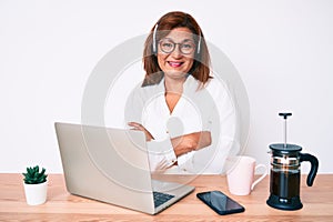 Middle age brunette hispanic woman working at the office wearing operator headset happy face smiling with crossed arms looking at