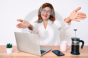 Middle age brunette hispanic woman working at the office wearing operator headset looking at the camera smiling with open arms for