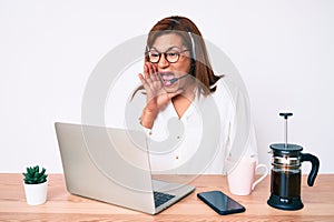 Middle age brunette hispanic woman working at the office wearing operator headset shouting and screaming loud to side with hand on