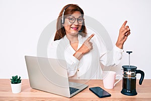 Middle age brunette hispanic woman working at the office wearing operator headset smiling and looking at the camera pointing with