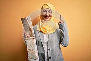 Middle age brunette tourist woman wearing traditional muslim hijab holding city map surprised with an idea or question pointing