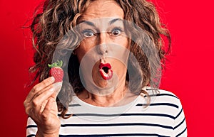 Middle age brunette woman eating healthy strawberry fruit over isolated red background scared and amazed with open mouth for