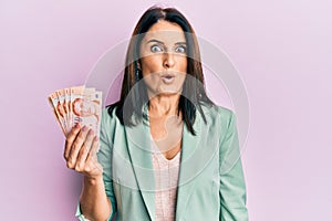 Middle age brunette woman holding 10 singapure dollars banknotes scared and amazed with open mouth for surprise, disbelief face