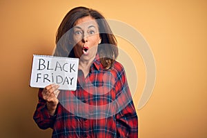 Middle age brunette woman holding black friday paper ad over isolated background scared in shock with a surprise face, afraid and