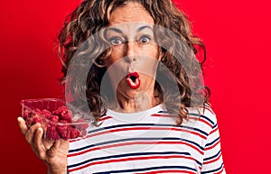Middle age brunette woman holding box of healthy raspberries over isolated red background scared and amazed with open mouth for