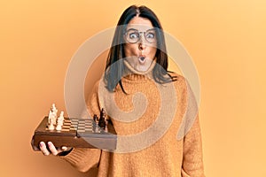 Middle age brunette woman holding chess board scared and amazed with open mouth for surprise, disbelief face