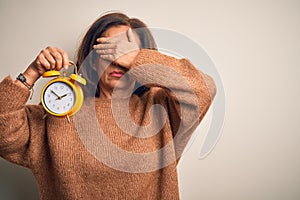 Middle age brunette woman holding clasic alarm clock over isolated background covering eyes with hand, looking serious and sad