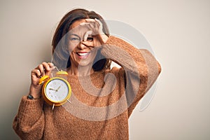 Middle age brunette woman holding clasic alarm clock over isolated background doing ok gesture with hand smiling, eye looking