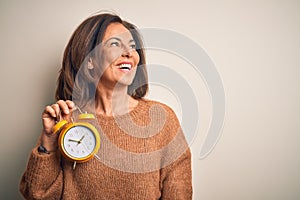 Middle age brunette woman holding clasic alarm clock over isolated background looking away to side with smile on face, natural