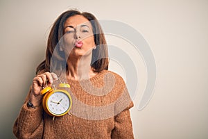 Middle age brunette woman holding clasic alarm clock over isolated background looking at the camera blowing a kiss on air being