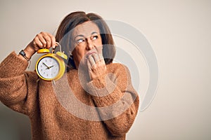 Middle age brunette woman holding clasic alarm clock over isolated background looking stressed and nervous with hands on mouth