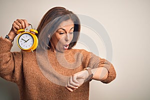 Middle age brunette woman holding clasic alarm clock over isolated background Looking at the watch time worried, afraid of getting