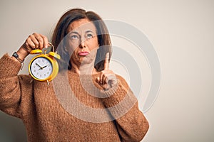 Middle age brunette woman holding clasic alarm clock over isolated background Pointing up looking sad and upset, indicating