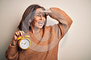Middle age brunette woman holding clasic alarm clock over isolated background very happy and smiling looking far away with hand