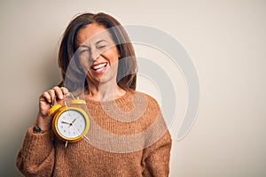 Middle age brunette woman holding clasic alarm clock over isolated background winking looking at the camera with sexy expression,