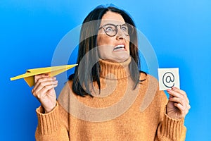 Middle age brunette woman holding email symbol and paper plane angry and mad screaming frustrated and furious, shouting with anger