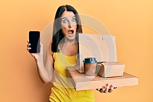 Middle age brunette woman holding take away food showing smartphone screen afraid and shocked with surprise and amazed expression,