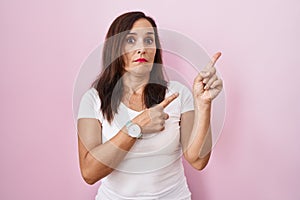 Middle age brunette woman standing over pink background pointing aside worried and nervous with both hands, concerned and