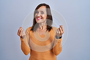 Middle age brunette woman standing wearing orange sweater doing money gesture with hands, asking for salary payment, millionaire