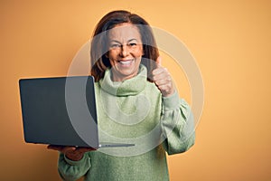 Middle age brunette woman using computer laptop over yellow background happy with big smile doing ok sign, thumb up with fingers,