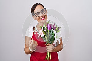 Middle age brunette woman wearing apron working at florist shop holding bouquet beckoning come here gesture with hand inviting