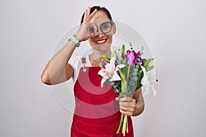 Middle age brunette woman wearing apron working at florist shop holding bouquet doing ok gesture with hand smiling, eye looking