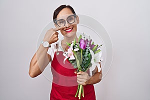 Middle age brunette woman wearing apron working at florist shop holding bouquet pointing with hand finger to face and nose,