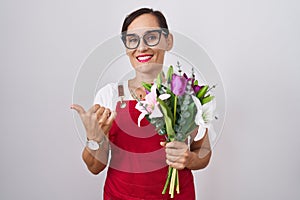 Middle age brunette woman wearing apron working at florist shop holding bouquet pointing to the back behind with hand and thumbs