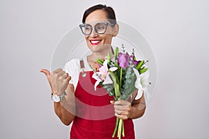 Middle age brunette woman wearing apron working at florist shop holding bouquet smiling with happy face looking and pointing to