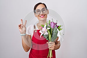 Middle age brunette woman wearing apron working at florist shop holding bouquet smiling with happy face winking at the camera