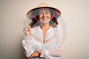 Middle age brunette woman wearing asian traditional conical hat over white background with a big smile on face, pointing with hand