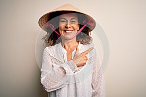 Middle age brunette woman wearing asian traditional conical hat over white background cheerful with a smile of face pointing with