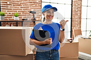 Middle age brunette woman wearing delivery uniform at house moving pointing thumb up to the side smiling happy with open mouth