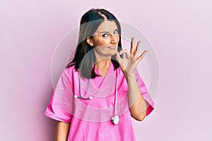 Middle age brunette woman wearing doctor uniform and stethoscope smiling positive doing ok sign with hand and fingers