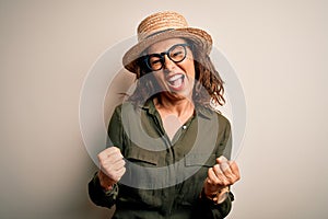 Middle age brunette woman wearing glasses and hat standing over isolated white background very happy and excited doing winner